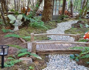 Eine Holzbrücke über einen kiesbestreuten Pfad in einem bewaldeten Gebiet mit Farnen und einer Statue einer Fee.