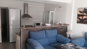 A modern living room with a blue sofa, a glass coffee table, and a kitchen area with a stainless steel refrigerator, microwave, and stove. - Family house in a quiet university neighborhood (Málaga)