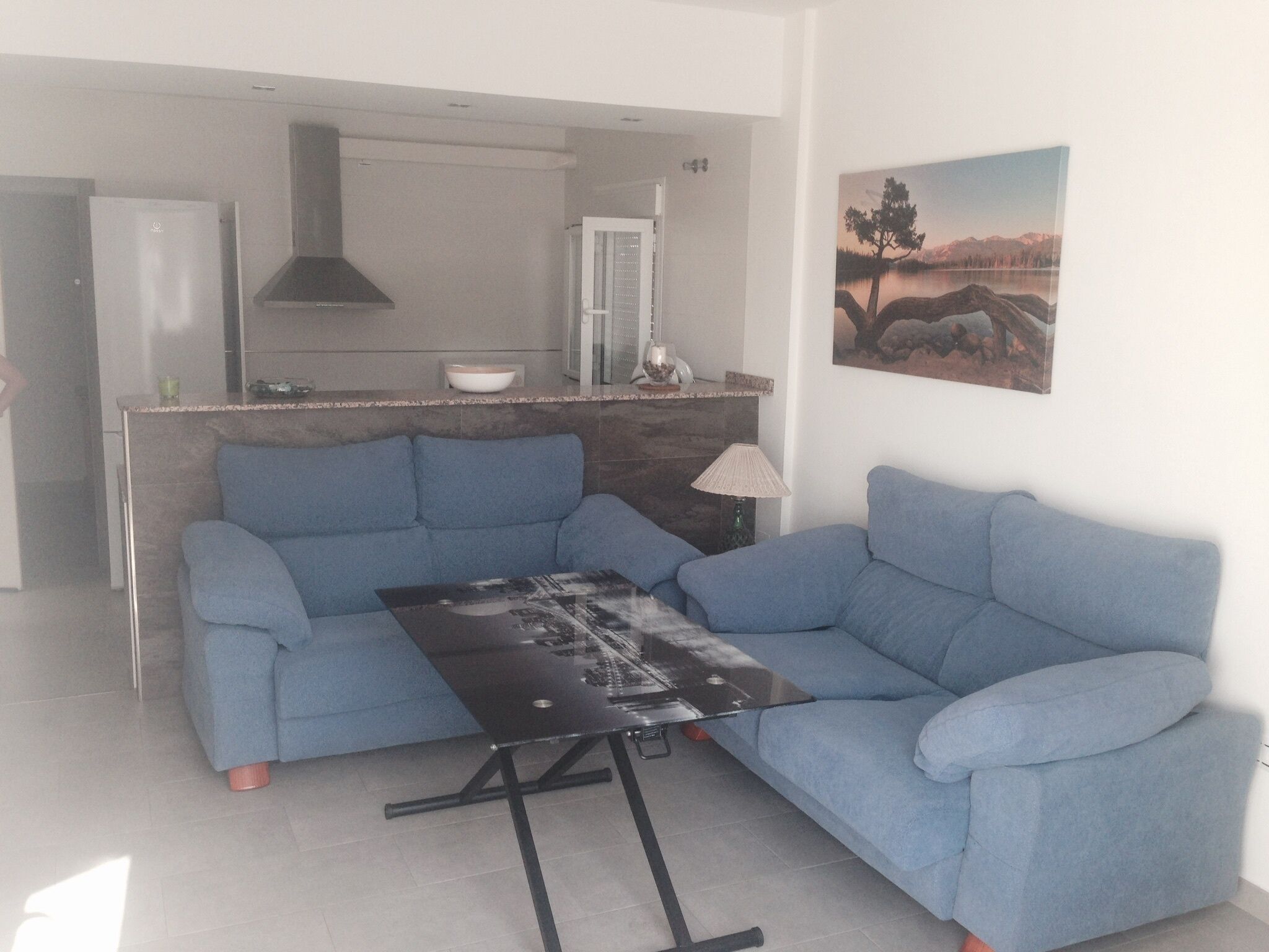 A modern living room with a blue sofa, a glass coffee table, and a kitchen area in the background.