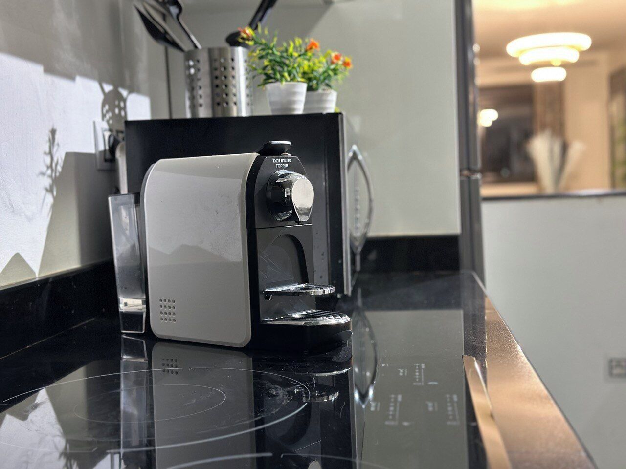 A coffee machine on a black countertop with a white base, a small potted plant, and a metal utensil holder in the background.