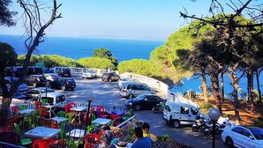 Eine Küstenstraße mit geparkten Autos und einem weißen Lieferwagen, mit Blick auf einen Essbereich mit bunten Stühlen und Tischen.