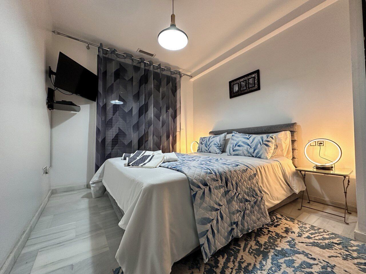 Een slaapkamer met een bed, een nachtkastje met een lamp, een aan de muur bevestigde televisie en een kamerkleed met dessin.