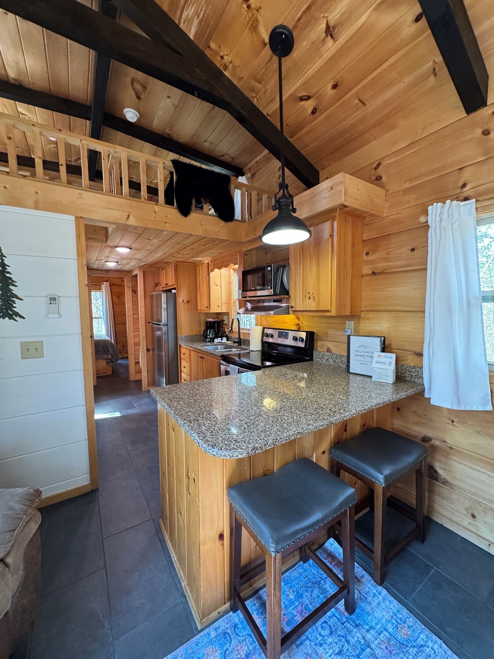 A wooden cabin interior with a kitchen area featuring granite countertops, a central island with bar stools, a hanging light fixture, and a welcome sign.