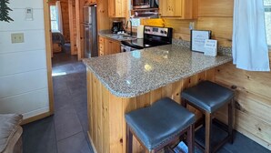 Interior de una cabaña de madera con zona de cocina que incluye encimeras de granito, una isla central con taburetes de bar, una lámpara colgante y un letrero de bienvenida.