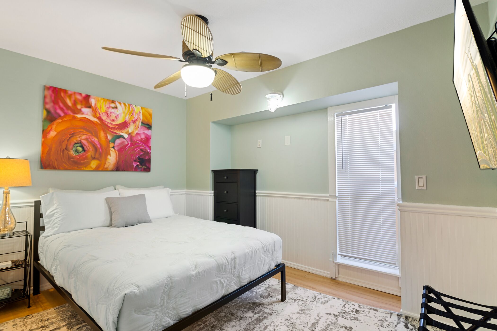 Quarto com cama, ventilador de teto, quadro, cômoda e janela com persianas.