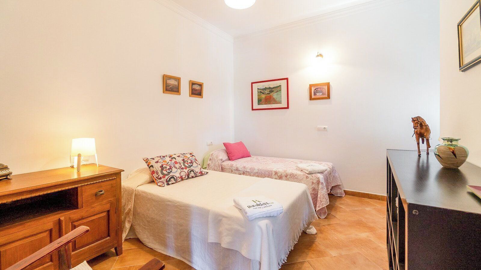 A bedroom with two beds, a desk, and a dresser. There are wall-mounted lights and framed pictures.