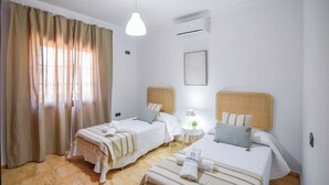 A hotel room with two beds, a window with curtains, an air conditioning unit, and a bedside table with a lamp.