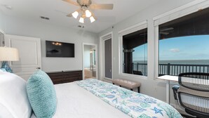 Un dormitorio con cama, ventilador de techo, televisor, ventana con vista al océano y una silla junto a la ventana.