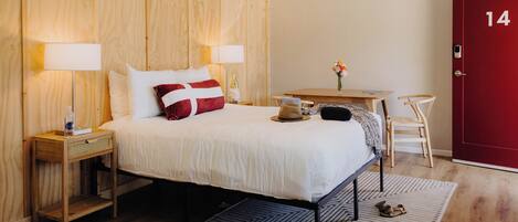 A hotel room with a wooden bed, two bedside tables, a desk with a chair, a red door, and a wooden floor.