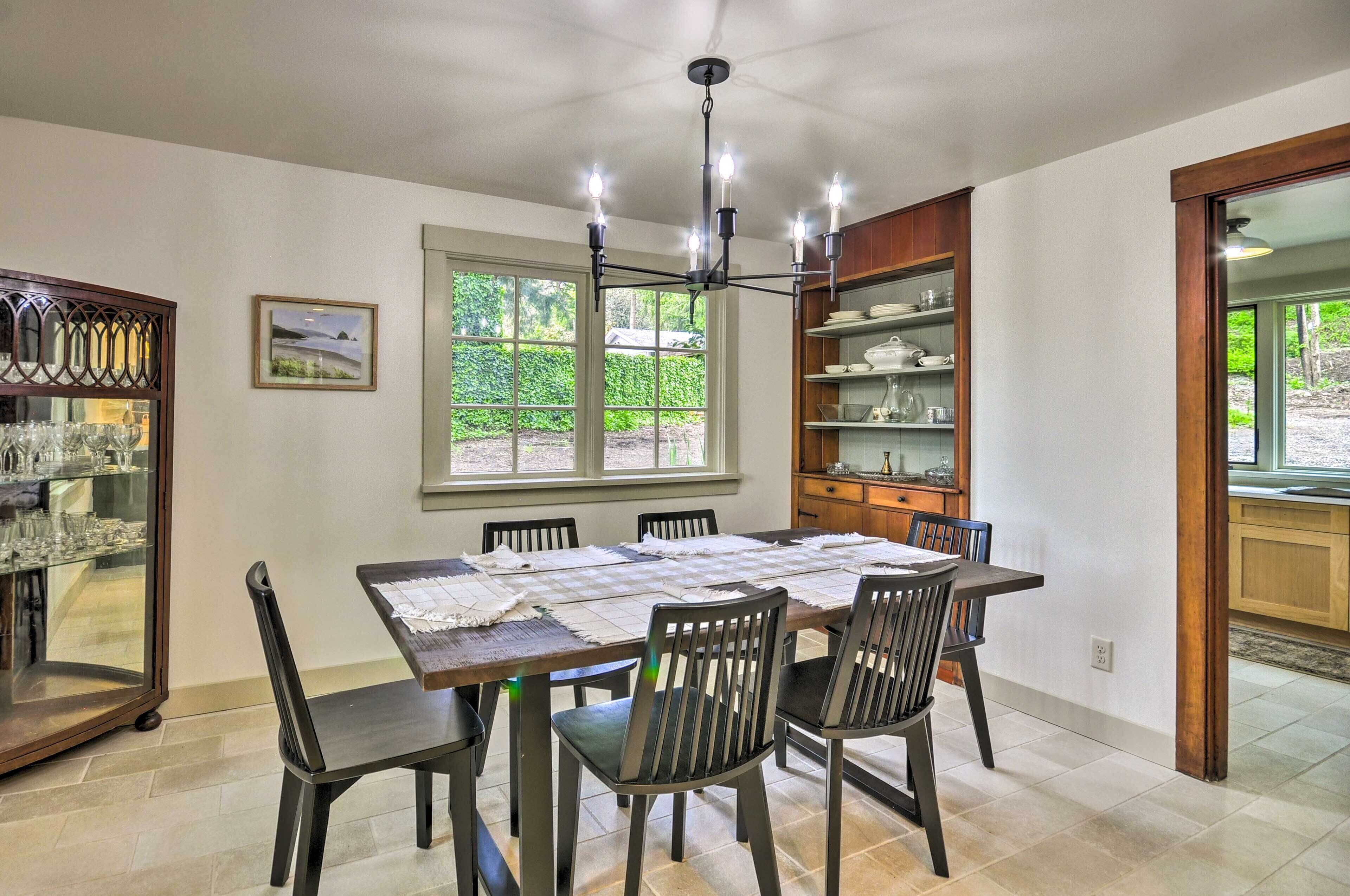 A dining area with a large table, six chairs, a chandelier, and a view of greenery outside.