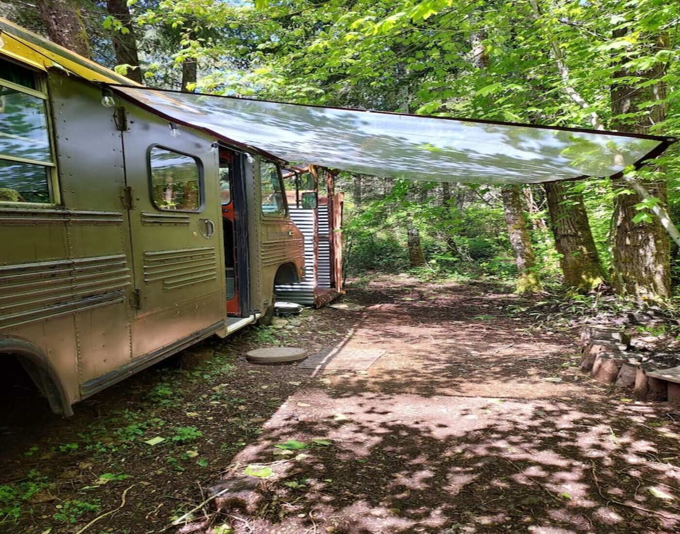 Une vieille caravane est garée dans une zone boisée, et un passage couvert mène à une structure en bois.