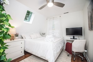 A bedroom with a bed, a desk, a chair, a television, a window, and a ceiling fan.