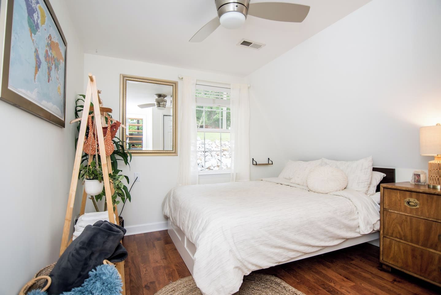 A bedroom with a bed, a dresser, a ceiling fan, a window with curtains, a plant, and a map on the wall.