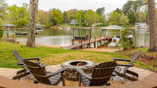 Un patio frente al lago con sillas Adirondack, una fogata y un muelle que se adentra en el agua.