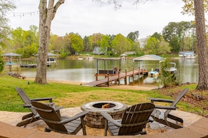 A lakeside patio with Adirondack chairs, a fire pit, and a dock extending into the water.