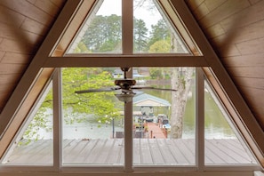 A room with a sloped ceiling and a view of a dock with boats and a house across the water.