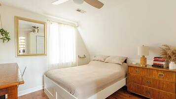 Une chambre à coucher comprenant un lit, une commode, un miroir, un ventilateur de plafond et une fenêtre avec des rideaux.