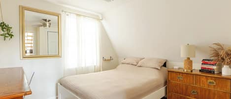 Une chambre à coucher comprenant un lit, une commode, un miroir, un ventilateur de plafond et une fenêtre avec des rideaux.
