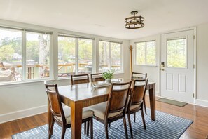 A dining area with a wooden table and chairs, large windows with a view of the outdoors, and a door with a doormat.