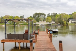 A wooden dock leading to a covered seating area with a view of a house and a boat on the water.