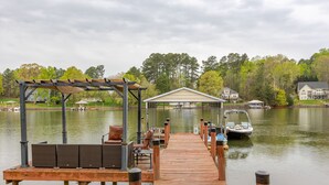 Un muelle de madera conduce a un área cubierta con sillas y una mesa, con vistas a un lago tranquilo y casas al fondo.