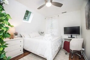 Une chambre à coucher comprenant un lit, un bureau, une chaise, un téléviseur, un ventilateur de plafond et une fenêtre avec des stores.