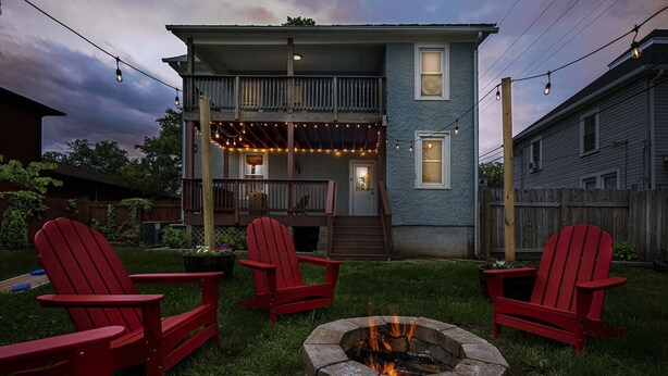 Een tweelaags huis met een veranda, een vuurplaats en rode Adirondack-stoelen.