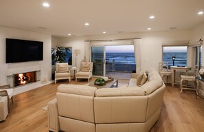 A spacious living room with a large beige sofa, a flat-screen TV mounted on the wall, a fireplace, and a view of the ocean through large sliding glass doors.