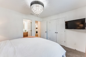 A bedroom with a large bed, a chandelier, white walls, a TV mounted on the wall, and a door leading to another room. - Luxury Atlanta Home(6 Bedrooms) (Atlanta)