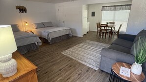 Ein gemütlicher Wohnbereich mit einer Couch, einem kleinen Tisch mit Pflanze, einem Essbereich mit Stühlen und zwei Betten mit Teppich.