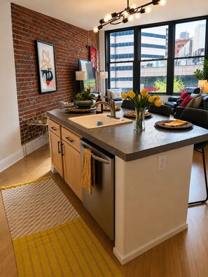 A kitchen with a large island, a brick wall, and a view of buildings through large windows.