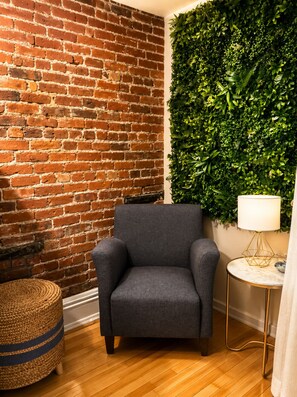 Une pièce avec un mur en briques, un fauteuil gris, une table ronde en bois surmontée d’une lampe dorée et un mur végétal vert.