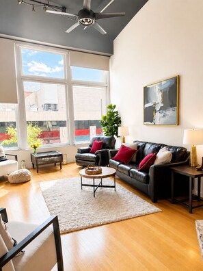 A modern living room with a black leather sofa, a round coffee table, a white rug, and a large window with a view of buildings.
