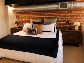 A bedroom with a brick wall, a bed with multiple pillows, a bedside table with a lamp, and a vent above the bed.