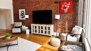 A living room with a brick wall, a flat-screen TV, white furniture, and a red floral painting. - NEW LISTING Charming 2/2 loft/heart of downtown. 5 min walk Convention Center (Denver)