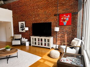 A living room with a brick wall, a flat-screen TV, white furniture, and a red floral painting.
