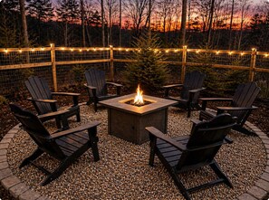 A fire pit surrounded by Adirondack chairs in a gravel area with a wooden fence and string lights.