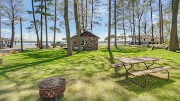 A stone cabin with a picnic table and a fire pit in a grassy area with tall trees and a lake in the background.
