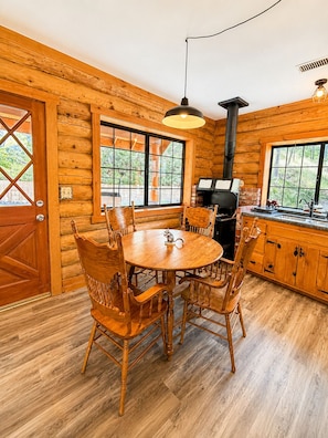 A wooden cabin interior with a round dining table, chairs, a built-in grill, and large windows.