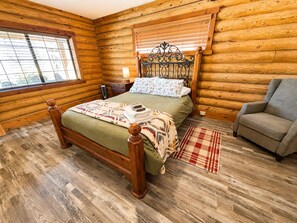 Een slaapkamer in een blokhut met een houten bed, een nachtkastje, een stoel en een raam.