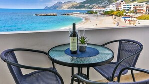 A balcony with a round table, two wicker chairs, a bottle of wine, and a small potted plant. The view includes a beach and buildings in the background.
