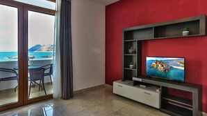 A modern living room with a large TV, a sliding glass door leading to a balcony with a view of the sea and mountains.