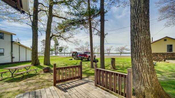 A wooden deck with picnic tables, a view of a lake, and a yellow building in the background. - Stone Lake Cabin w/ Private Deck & Fire Pit! (Stone Lake)