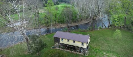 Een huis met veranda, omgeven door grasvelden en bomen, met een rivier en bergen op de achtergrond.