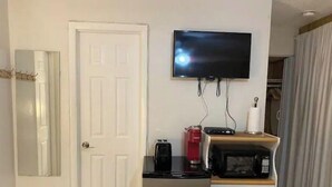 Habitación de hotel con una puerta blanca, un televisor de pantalla plana montado en la pared, cafetera, microondas y un mueble pequeño.