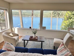 A room with a sofa, chairs, a table, and a view of a lake through large windows. - Beautiful Liberty Lake Waterfront House (Liberty Lake)