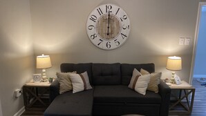 Uma sala de estar com um sofá seccional, duas mesas laterais com luminárias e um relógio de parede grande.