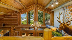 A log cabin interior with a large window, wooden ceiling, and a deer head decoration.