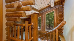 A log cabin interior with a wooden staircase and large windows overlooking a forest.