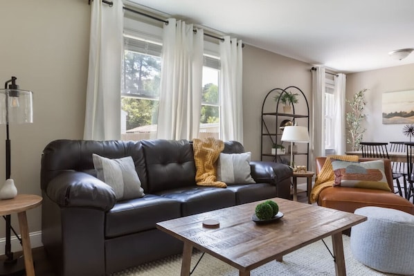 A living room with a dark leather sofa, a wooden coffee table, and a dining area with a table and chairs. - 3BR Oasis | Mins from Downtown - King Bed (Garner)